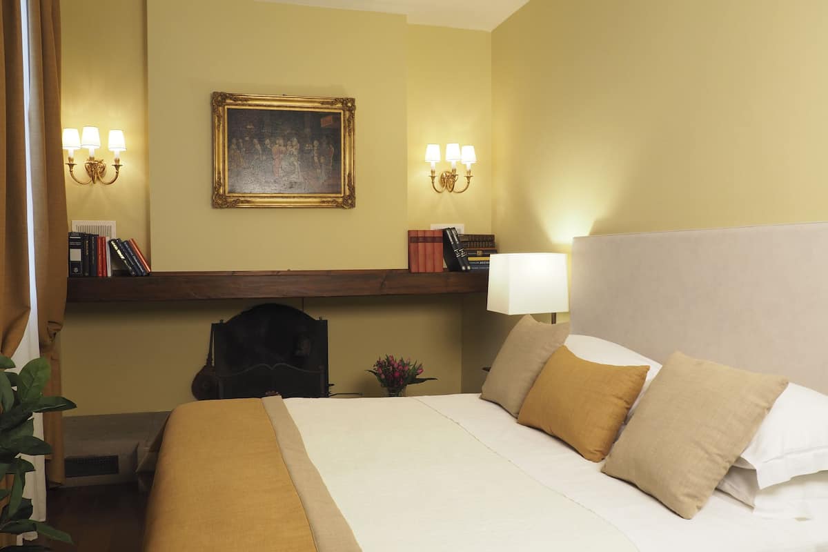 Quarto de hotel com uma cama grande, cabeceira, mesa de cabeceira, luminária, pintura na parede e prateleira com livros e objetos decorativos.