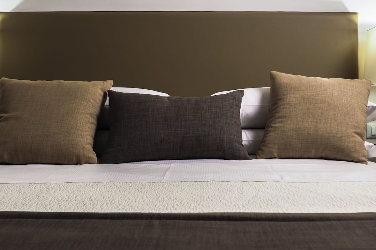 Uma cama com cabeceira, três almofadas e um edredom branco texturizado.