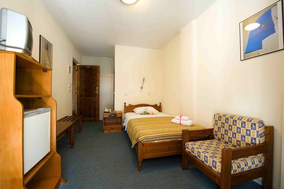 Una habitación pequeña y ordenada con una cama, una silla, un escritorio con una computadora y un refrigerador pequeño.