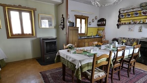 A dining room with a table set for four, wooden chairs, a patterned rug, and a kitchen area with a microwave and sink. - B&B Marie Thérèse (Torreano)