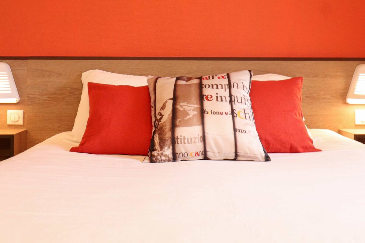 A hotel room with a bed, red pillows, a framed artwork on the wall, and bedside lamps.
