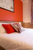 Un lit bien fait, avec une literie blanche et deux oreillers rouges, une œuvre d’art encadrée au mur et une tête de lit en bois.