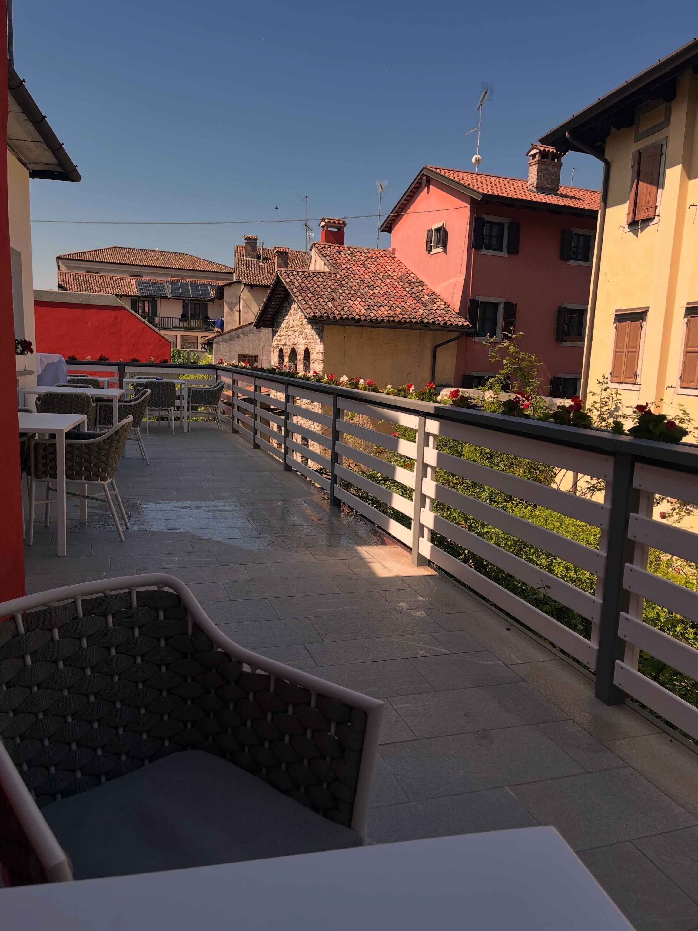 A terrace with outdoor seating, a white railing, and buildings with red-tiled roofs in the background.