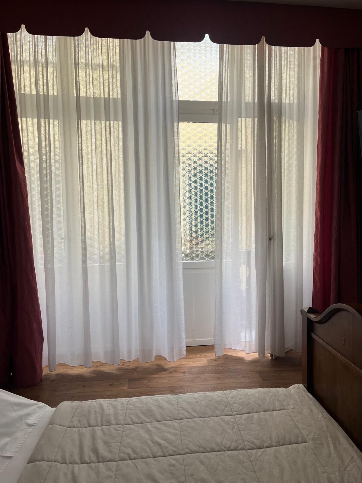 A bedroom with a bed, wooden floor, and a window with sheer curtains.