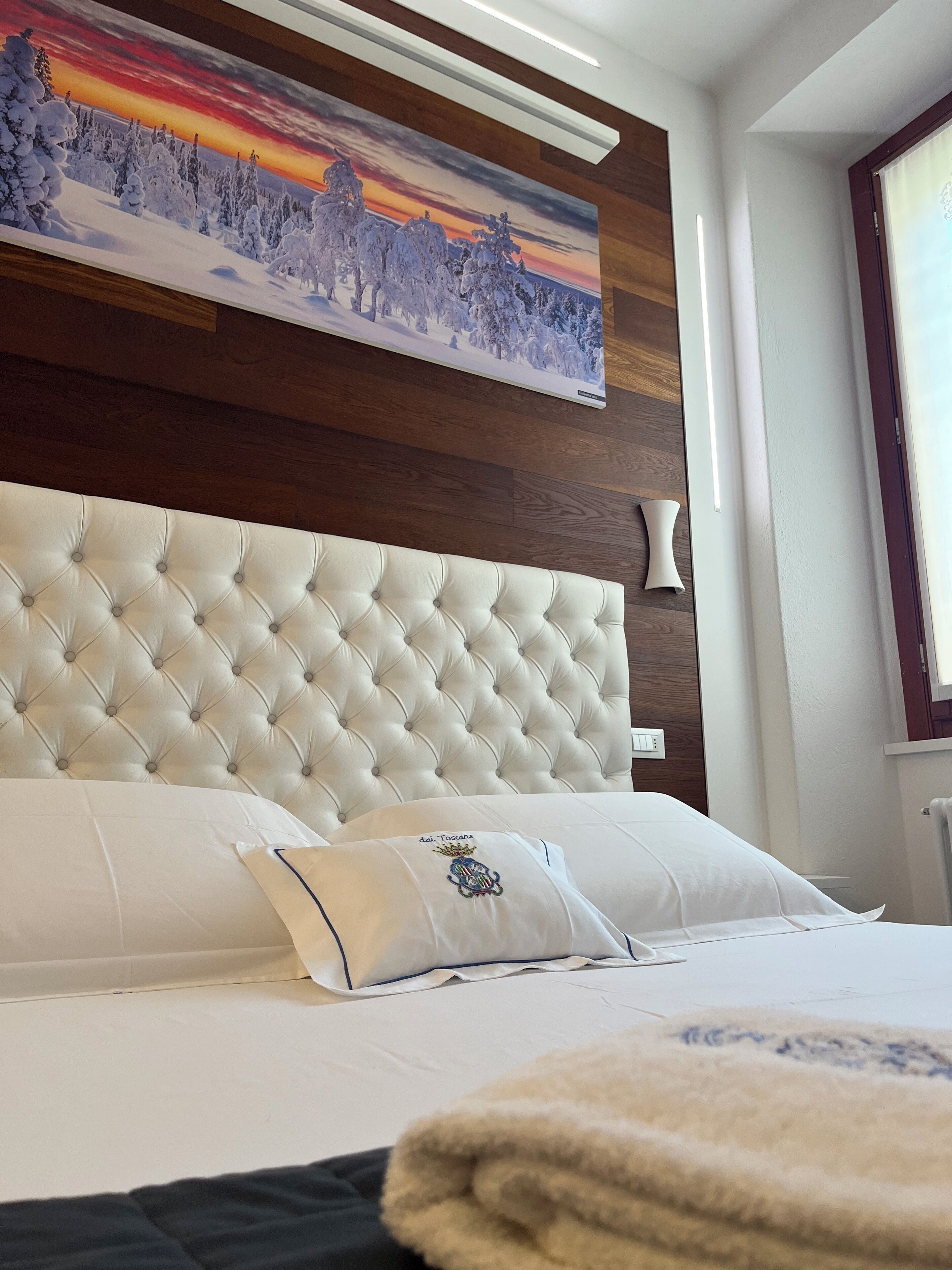 A bedroom with a tufted headboard, a painting of a snowy landscape, and a window with a view.