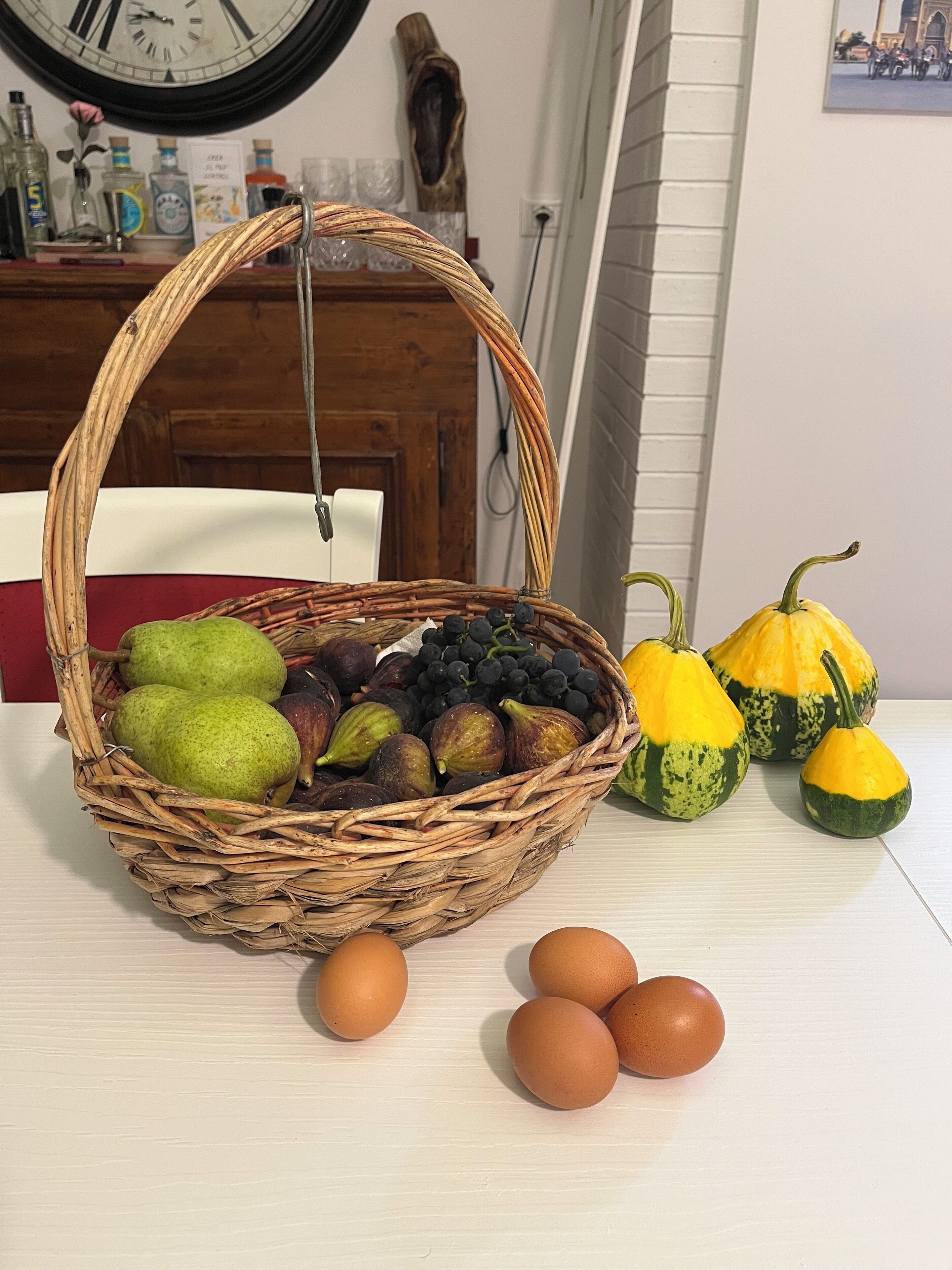 Ein Korankorb mit Obst und Gemüse, darunter Birnen, Feigen und Kürbis, auf einem weißen Tisch.