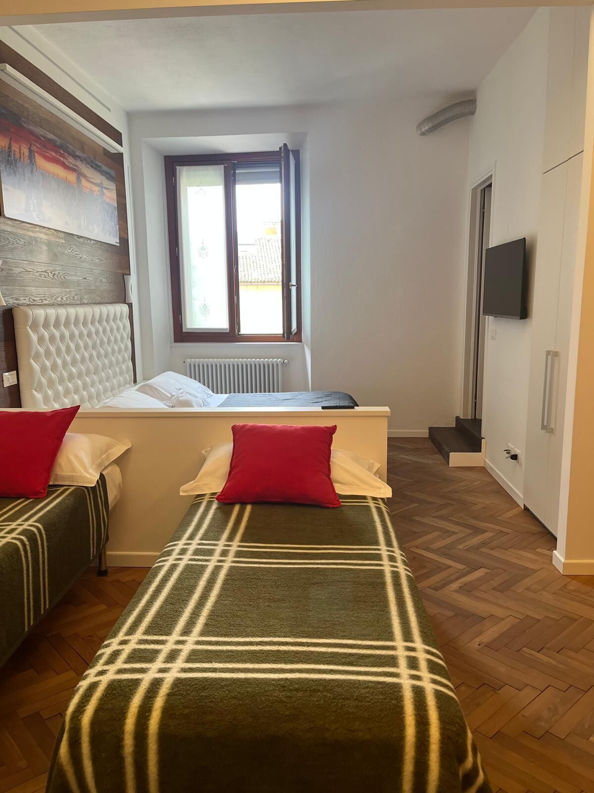 Un dormitorio con cama, dos mesitas de noche, una ventana, un radiador, televisor y suelo con patrón de espiga.