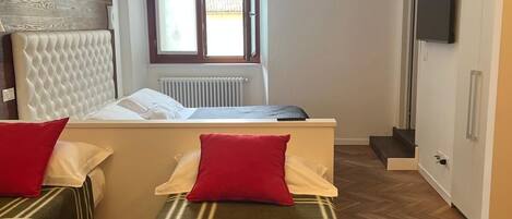 Un dormitorio con cama, dos mesitas de noche, una ventana, un radiador, televisor y suelo con patrón de espiga.