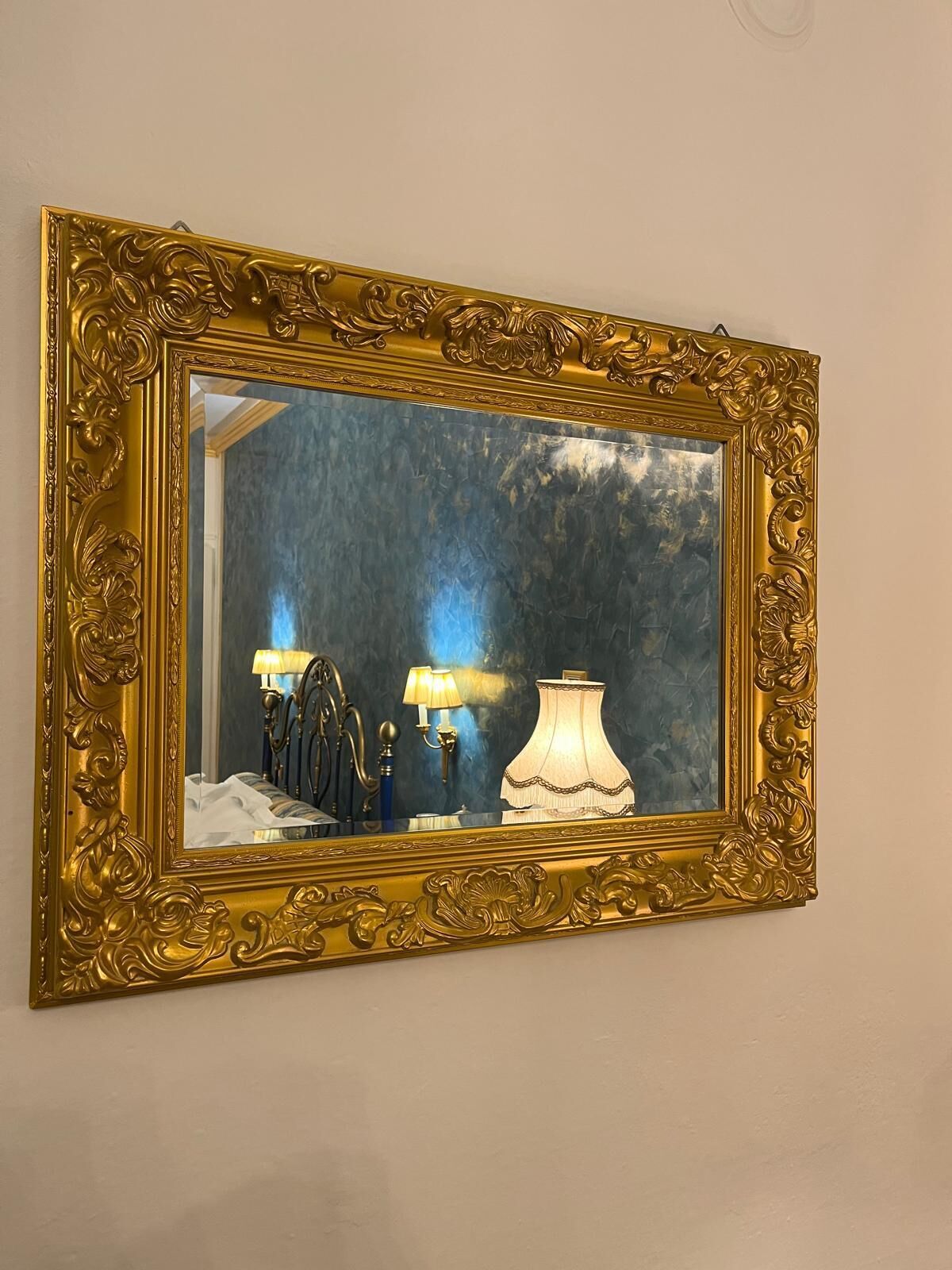 A mirror with an ornate gold frame reflecting a room with a bed, two lamps, and a wall with a textured pattern.