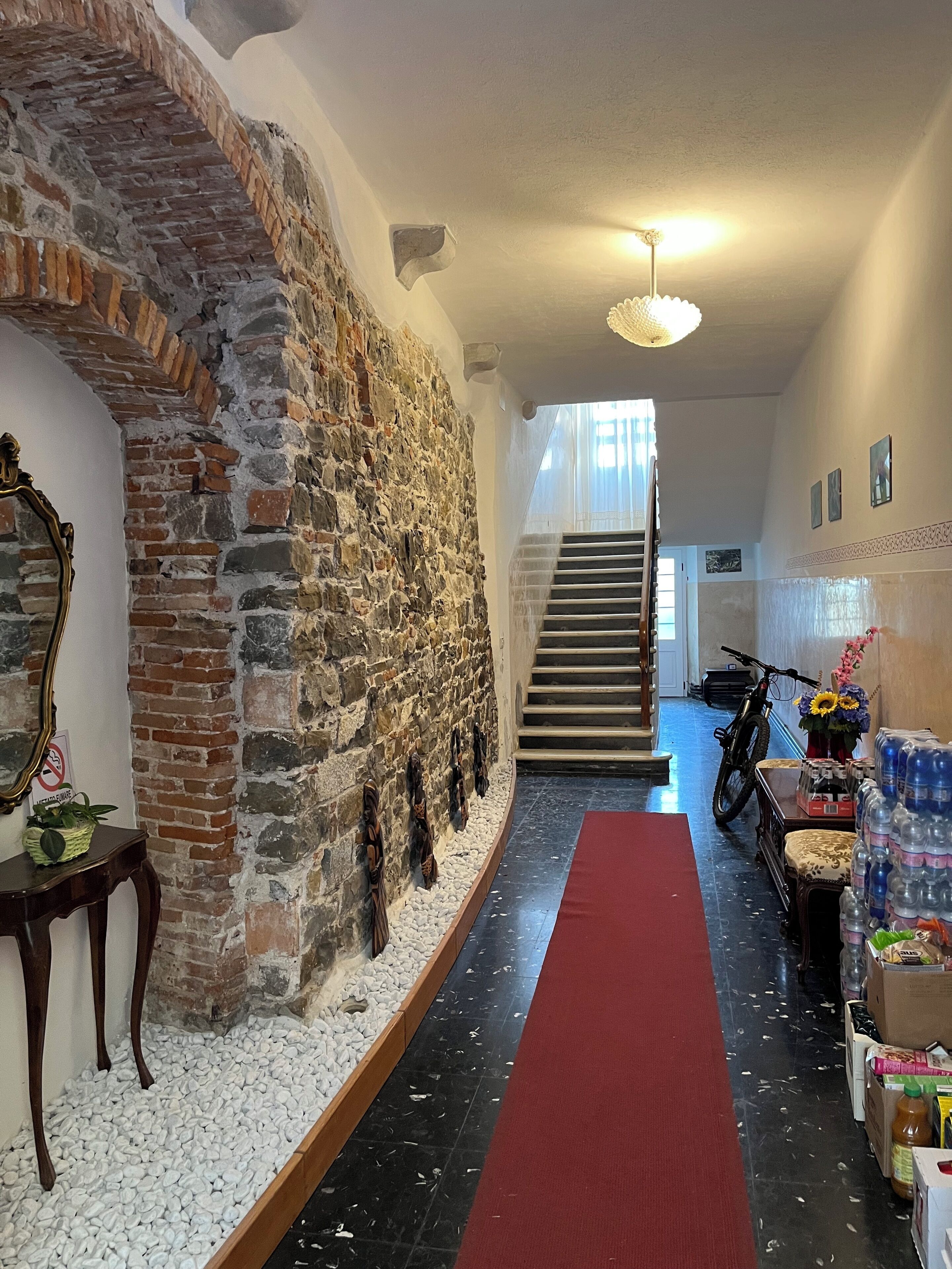 Un pasillo con pared de piedra, una mesa de madera con una cesta, alfombra roja y una escalera al fondo.