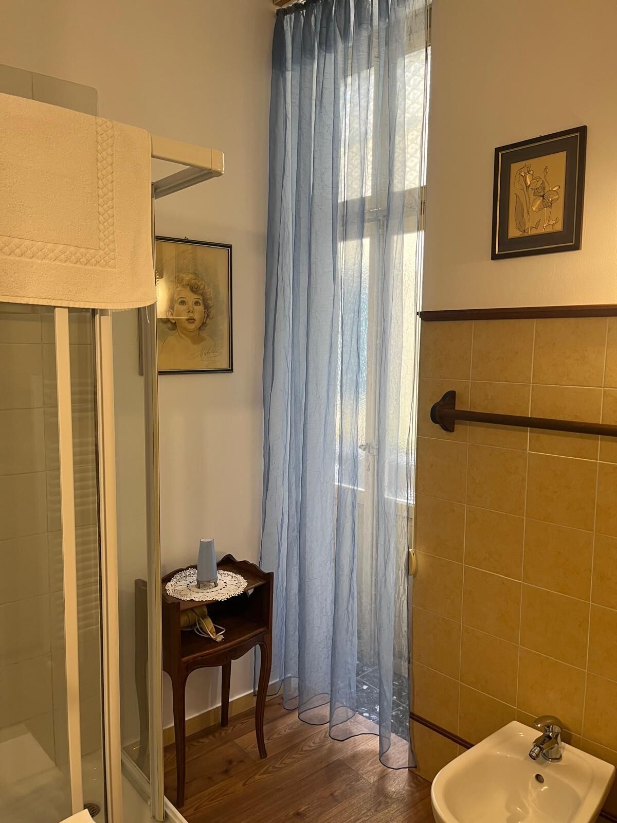 A bathroom with a shower, a sink, a framed picture, and a window with a sheer curtain.