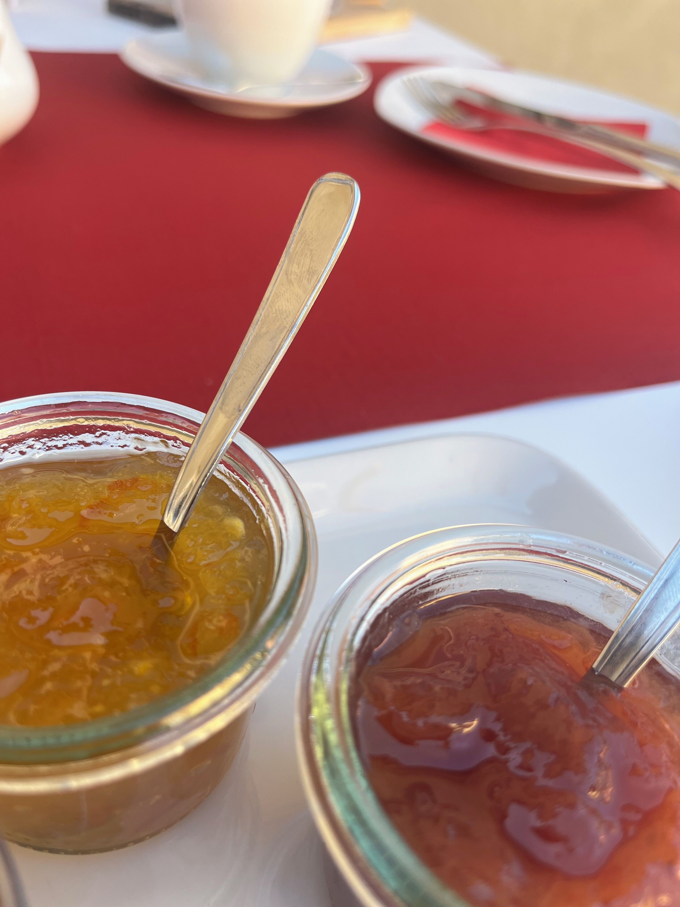 Two jars of jam on a white tray with a spoon in each, placed on a red tablecloth.