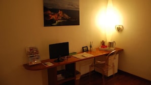Un petit bureau avec un bureau, un ordinateur et une chaise. Il y a un tableau au mur et une lampe sur le bureau.