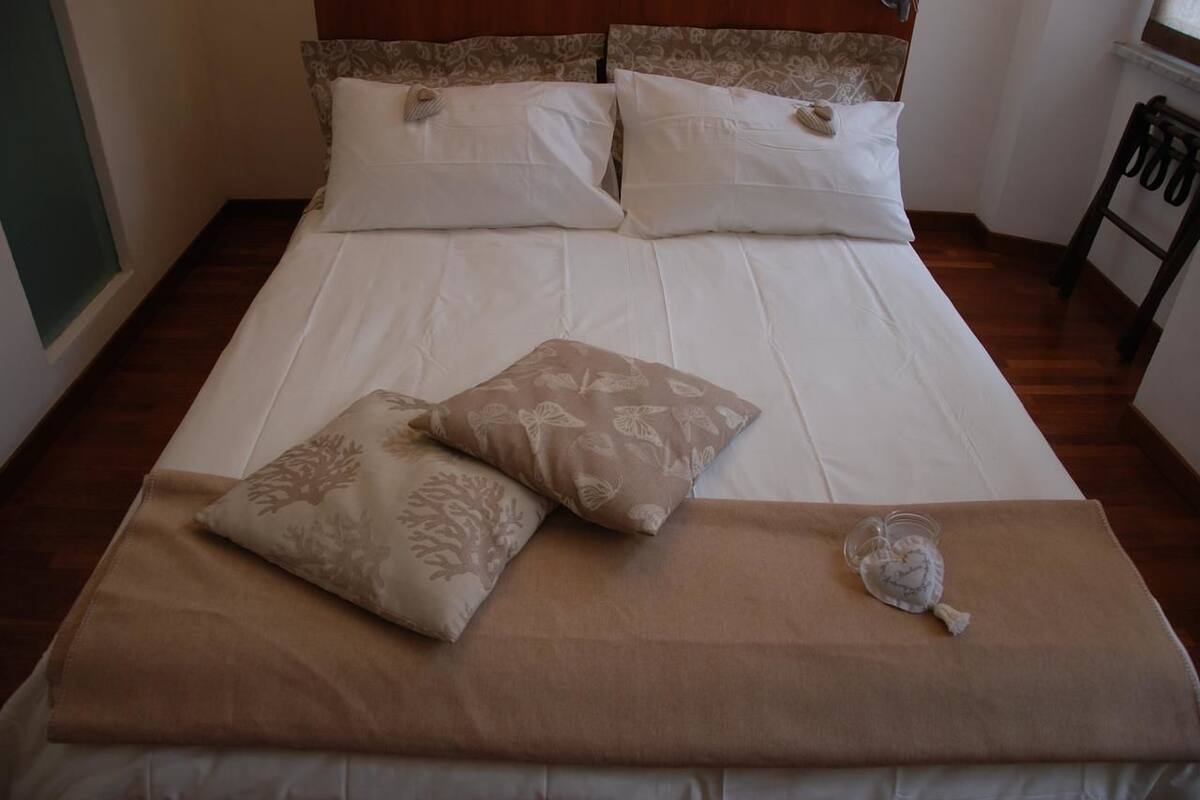 Ein ordentlich bezogenes Bett mit weißer und beiger Bettwäsche, zwei Zierkissen und ein kleines kissenförmiges Herz auf Holzdielen.