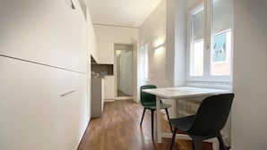 Une cuisine de taille réduite avec des meubles blancs, une table à manger avec deux chaises et une fenêtre équipée de stores.