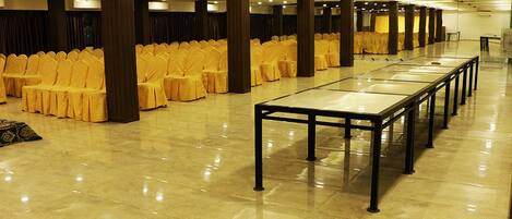 Een ruime zaal met rijen gele stoelen en een lange tafel, waarschijnlijk voor evenementen.