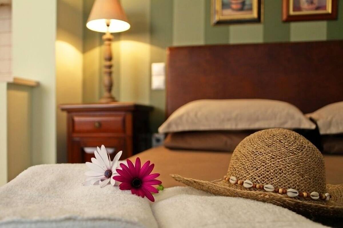 A neatly made bed with a white towel, a straw hat, and a pink flower.