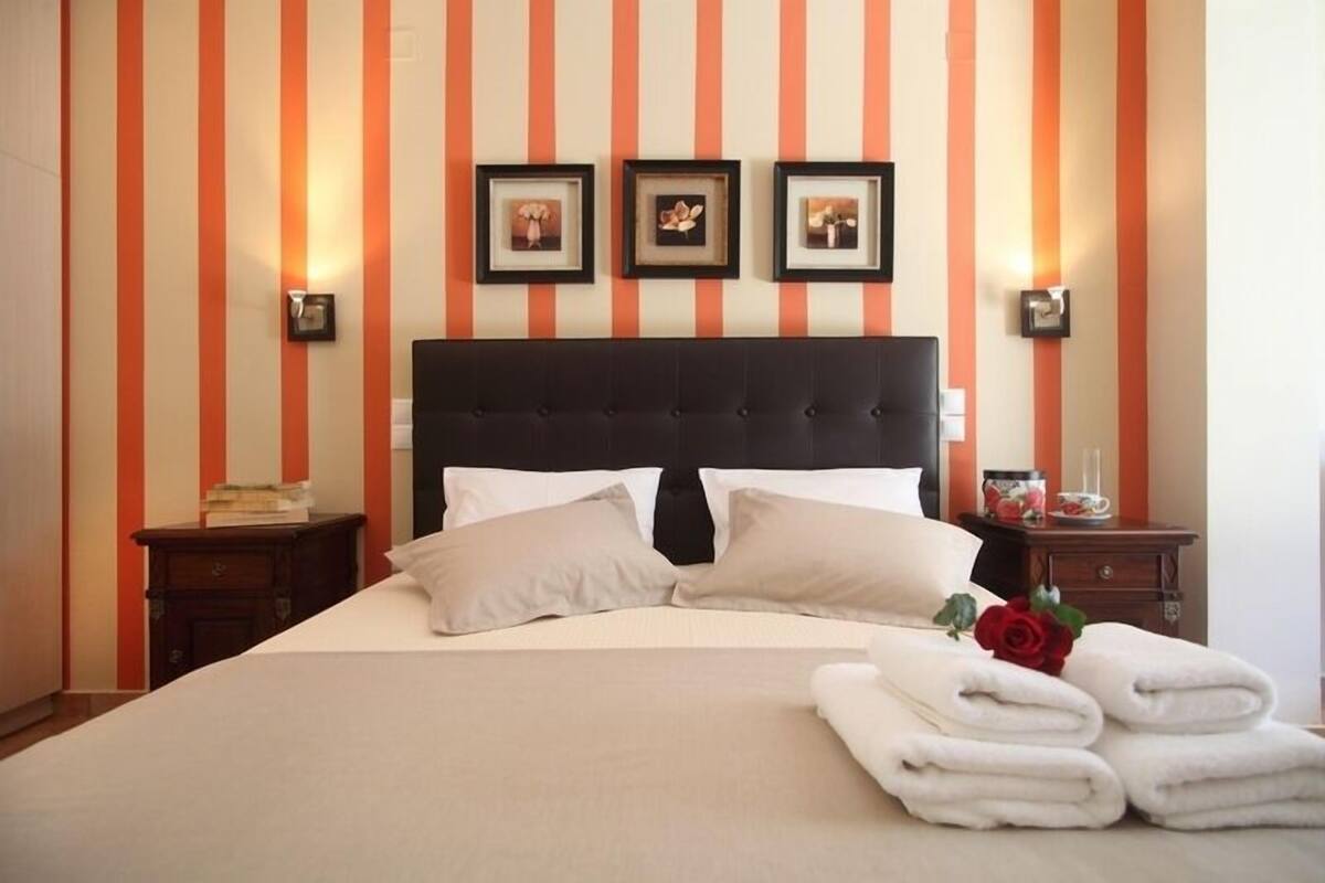 A neatly made bed with white linens and two pillows, a wooden headboard, and a nightstand with a lamp and a framed picture.