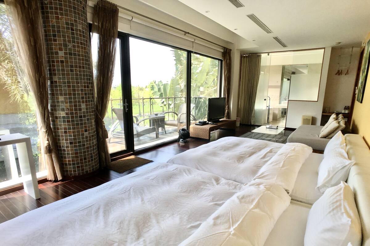 A large bed with white linens, a television on a small table, and a balcony with outdoor furniture.