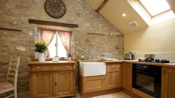 A kitchen with wooden cabinets, a stone wall, a clock, and a skylight. - The Cornmill Stable Cottages (York)