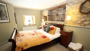 A bedroom with a wooden bed, a stone wall, a window, and a nightstand with a lamp. - The Cornmill Stable Cottages (York)