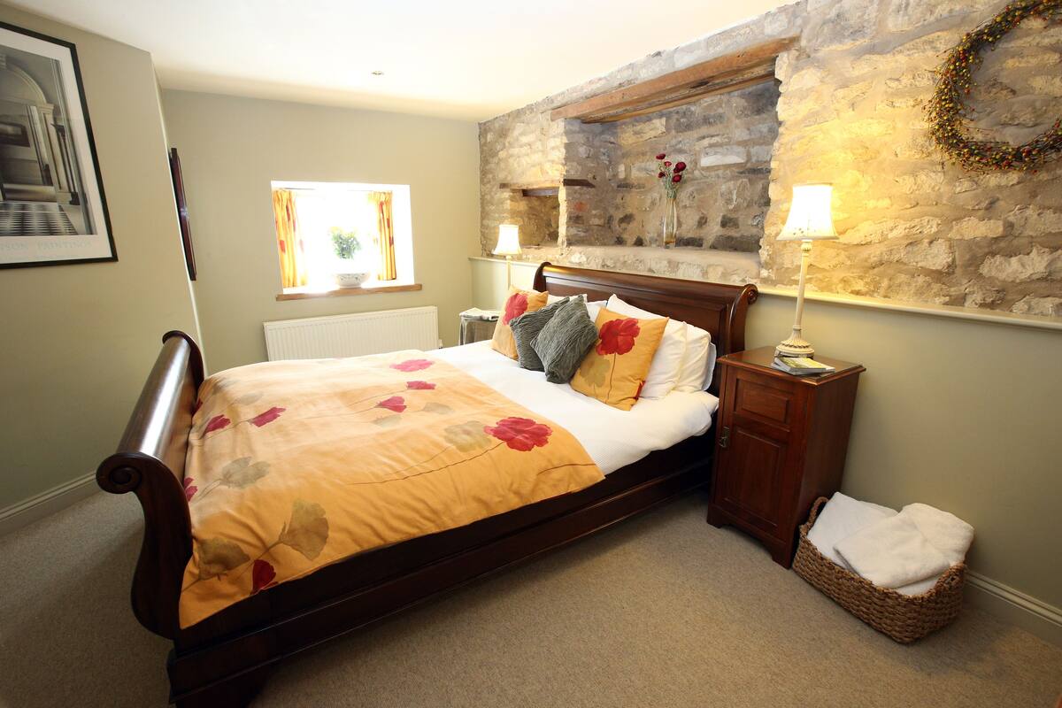 A bedroom with a wooden bed, a stone wall, a window, and a nightstand with a lamp.