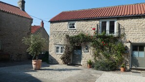 A stone building with a red-tiled roof, a small balcony with red flowers, and a gravel courtyard. - The Cornmill Stable Cottages (York)