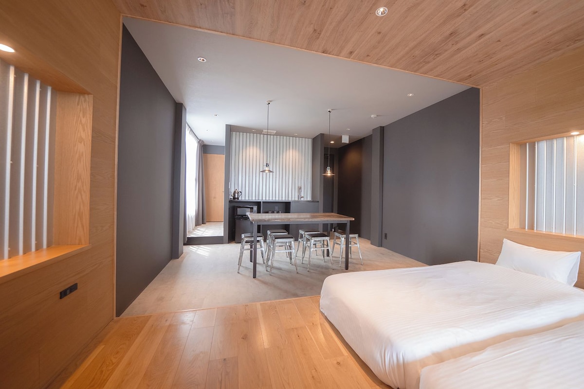 A modern hotel room with a wooden floor, a large bed, a desk with chairs, and a kitchenette in the background.
