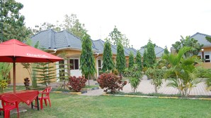 A courtyard with a red Coca-Cola branded umbrella, a table with red chairs, and a variety of plants and trees. - Country Motel Koboko (Koboko)