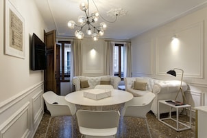 A modern living room with a round table, white sofas, a chandelier, and a flat-screen TV mounted on the wall.