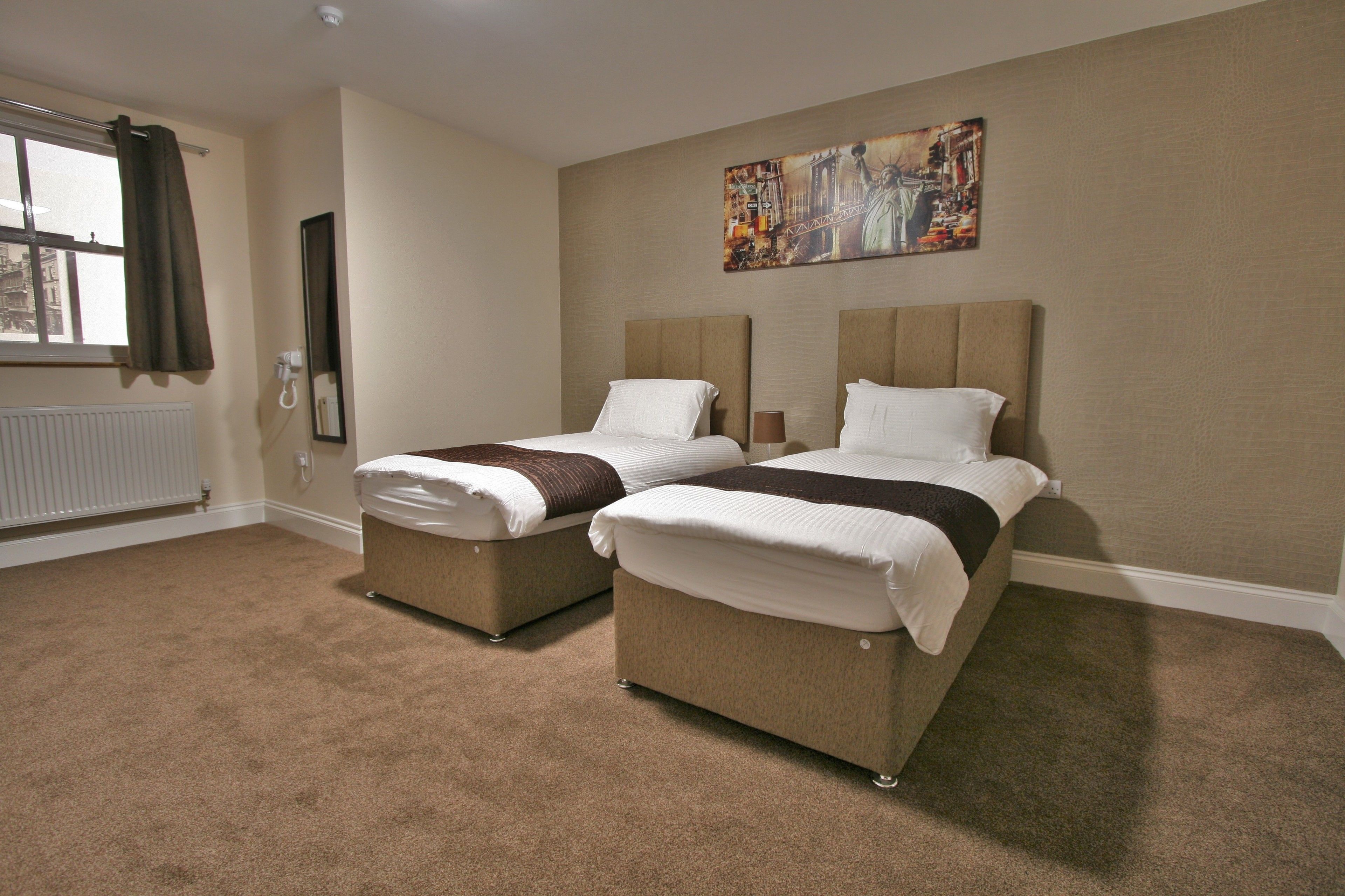 A hotel room with two beds, a radiator, a window with curtains, and a painting on the wall.