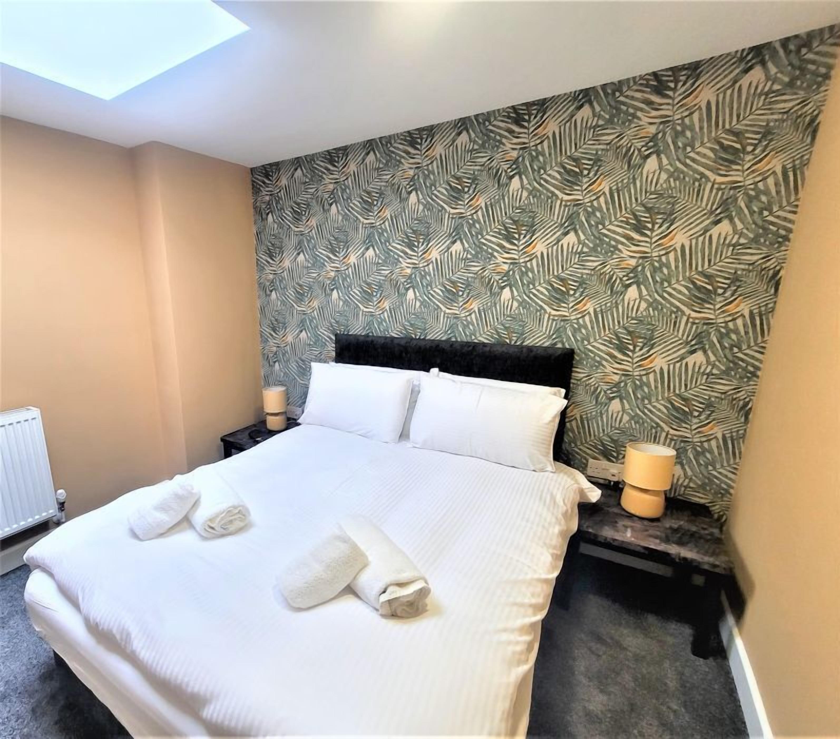 A neatly made bed with white linens and towels, a patterned wall, and a skylight above.