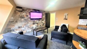 Una sala de estar con pared de piedra, un televisor de pantalla plana, dos sofás, una mesa de centro y un área de cocina con cubierta de madera y horno empotrado.