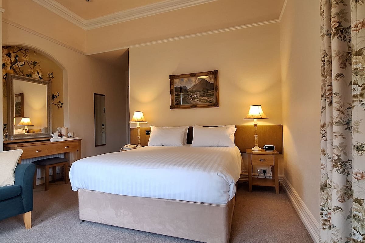 A hotel room with a bed, two bedside tables with lamps, a desk with a chair, a mirror, and floral curtains.