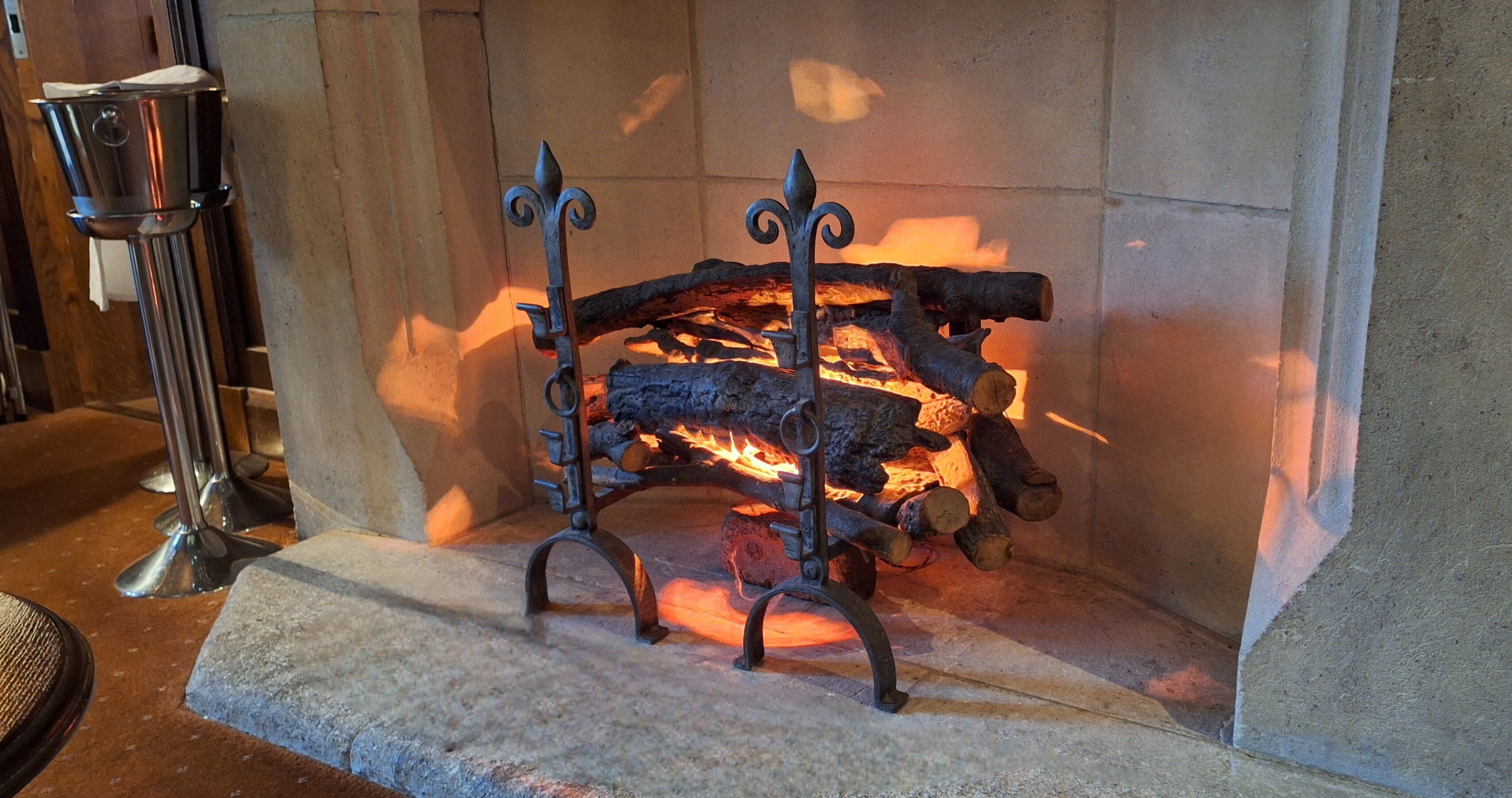 A fireplace with logs burning, a metal stand, and a metal container on the left.