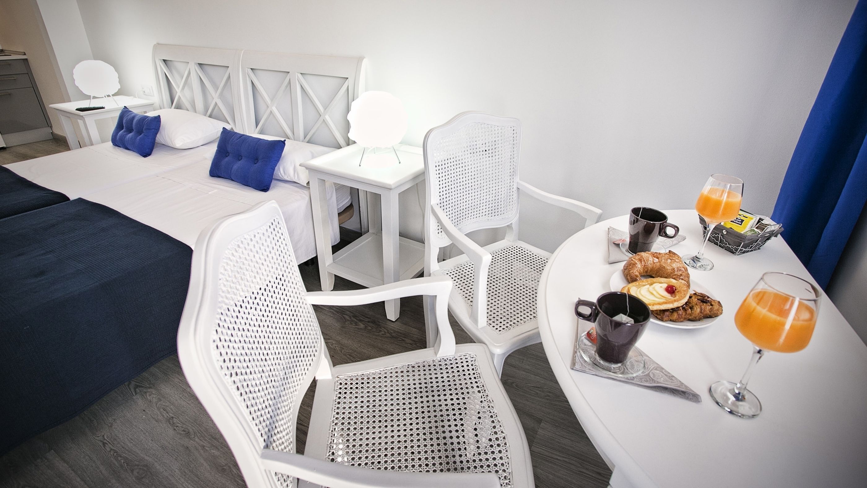 Habitación de hotel con cama, dos sillas blancas, una mesita con provisiones para el desayuno y una mesita de noche con lámpara.