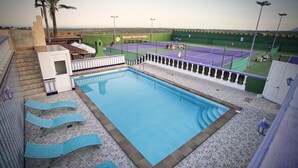 A swimming pool with lounge chairs, a tennis court, and a view of the city. - Corralejo Lodge (La Oliva)