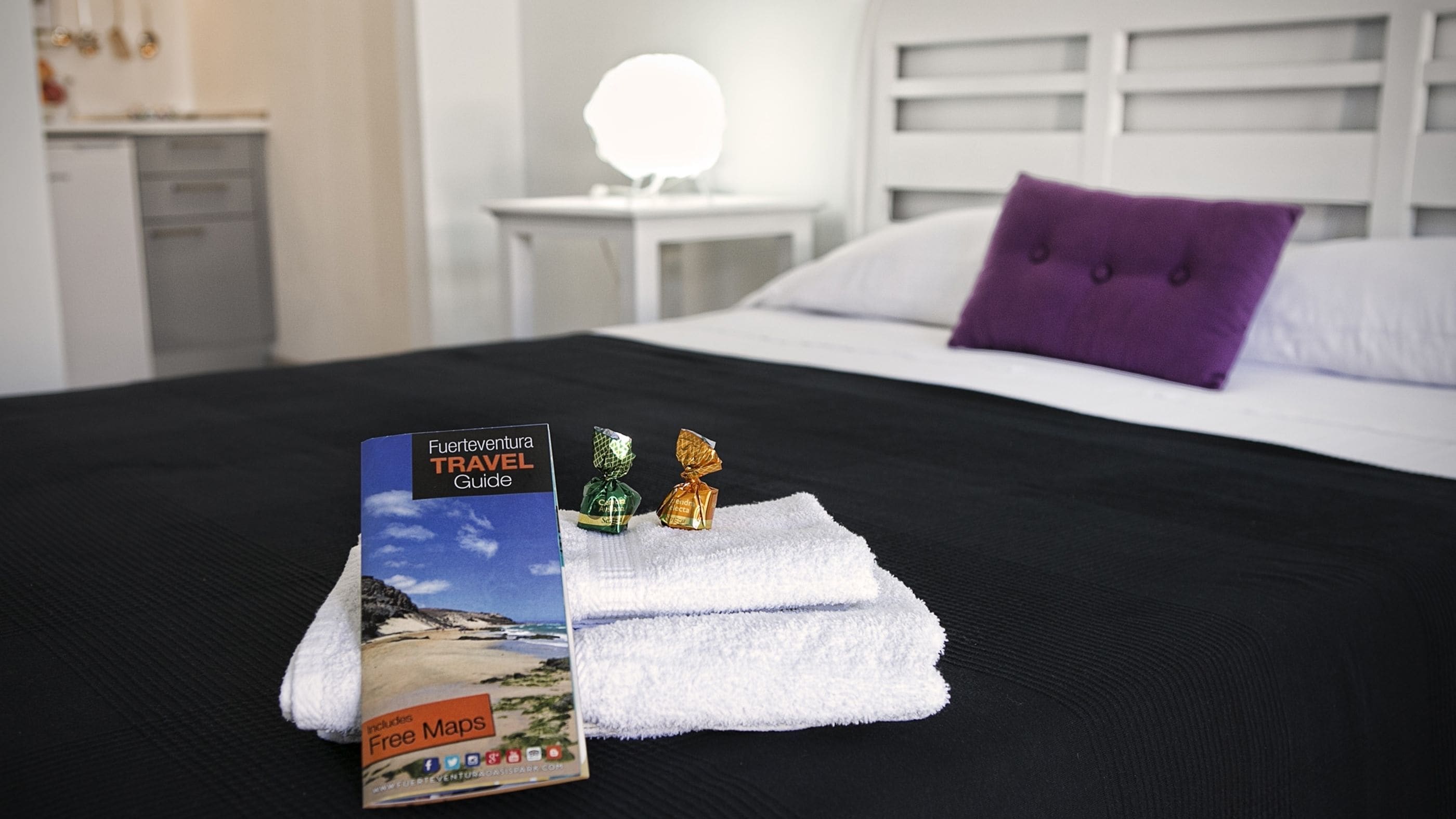 A neatly made bed with a travel guide, two wrapped chocolates, and a white towel on a black bedspread in a hotel room.
