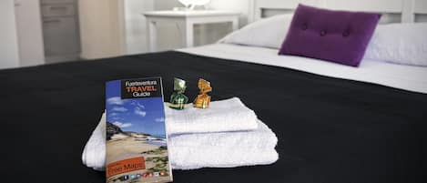 Un lit bien fait, avec un guide de voyage, deux chocolats emballés et une serviette blanche posée sur un couvre-lit noir, dans une chambre d’hôtel.