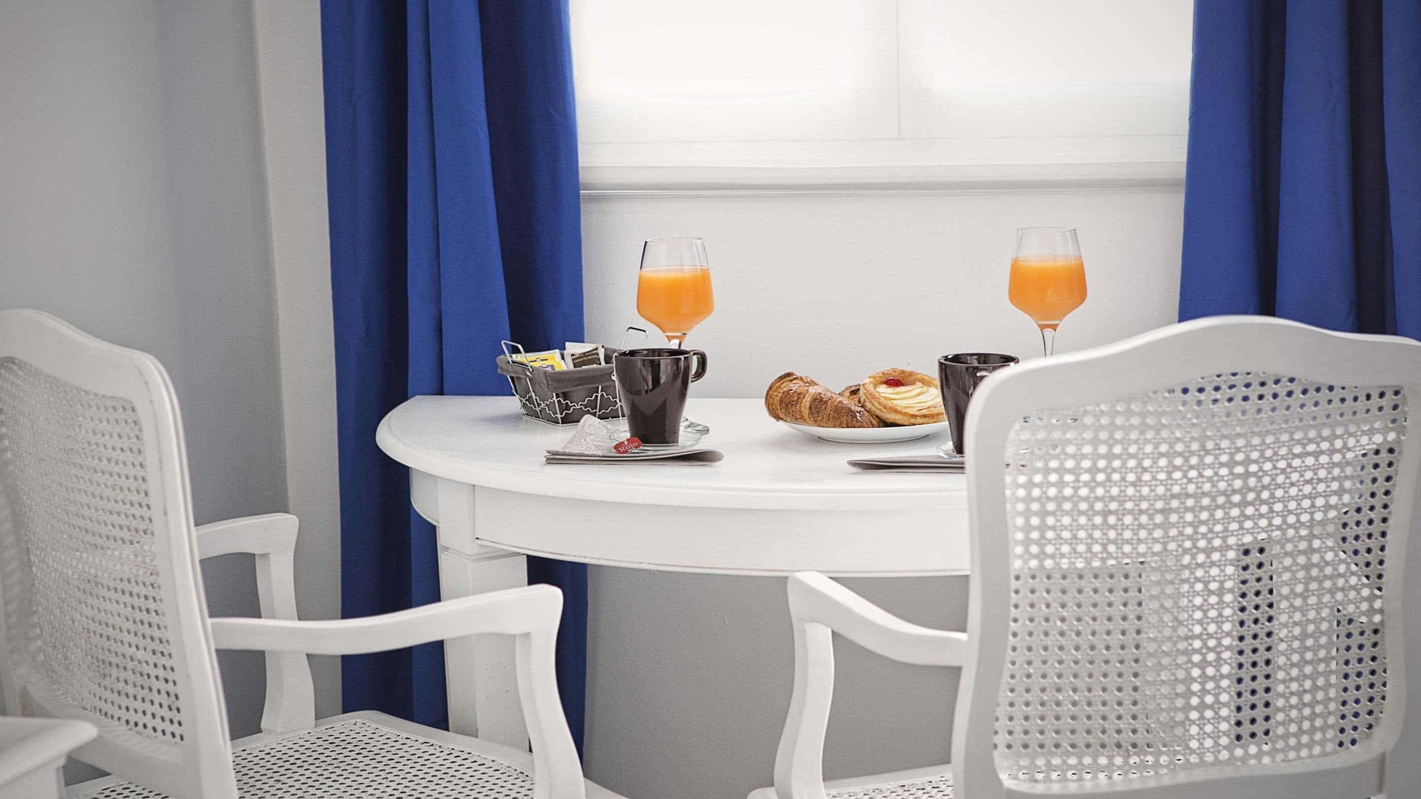 A breakfast table with two glasses of orange juice, a croissant, a muffin, and a basket of newspapers.