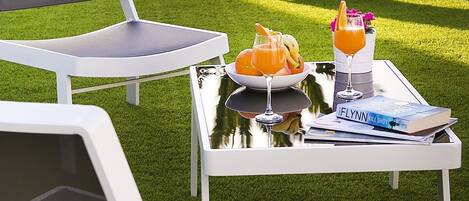 A patio with white lounge chairs, a table with drinks and a book, and a palm tree.