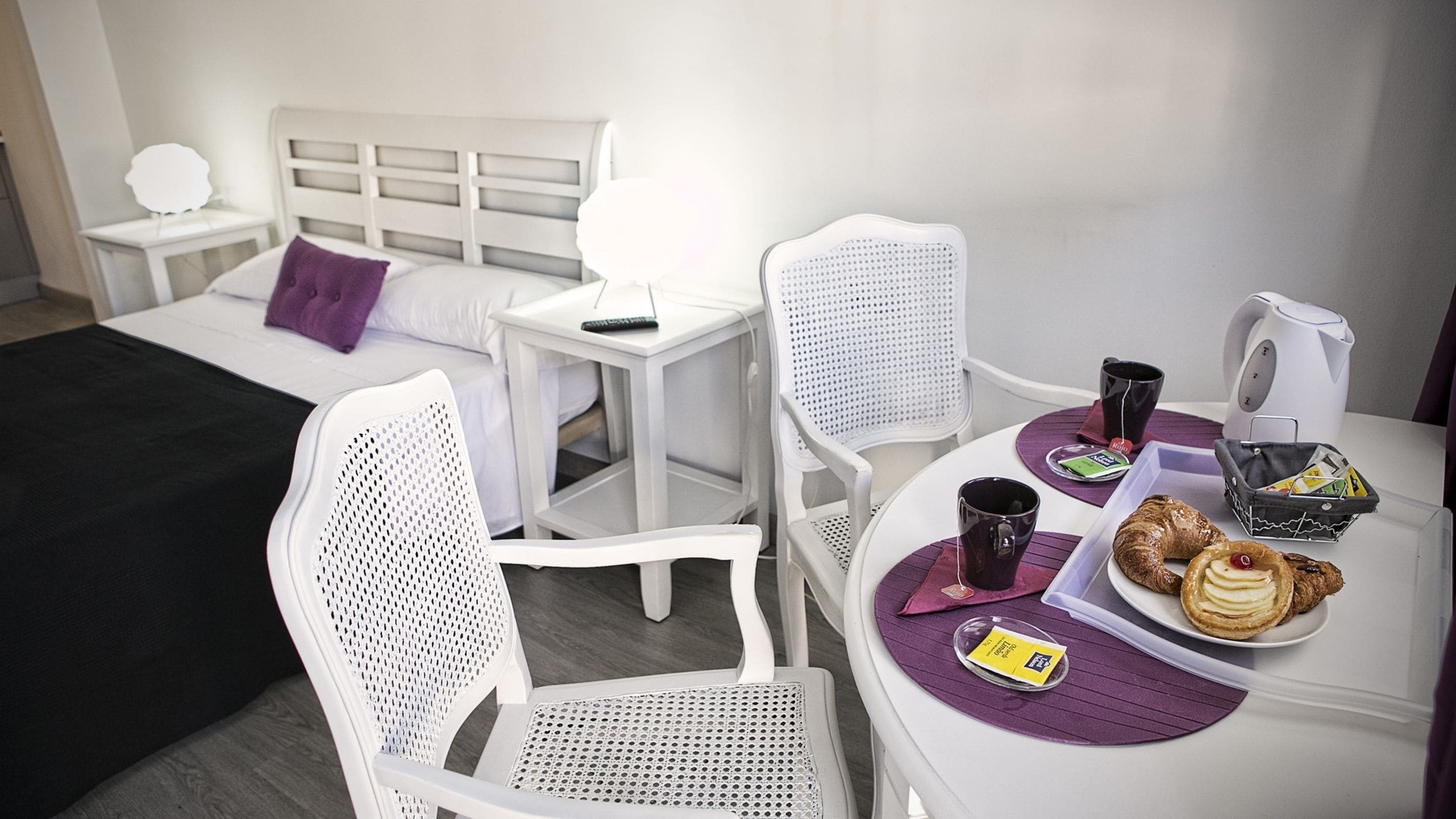Quarto de hotel com cama, escrivaninha, duas cadeiras, mesa com itens de café da manhã e chaleira.