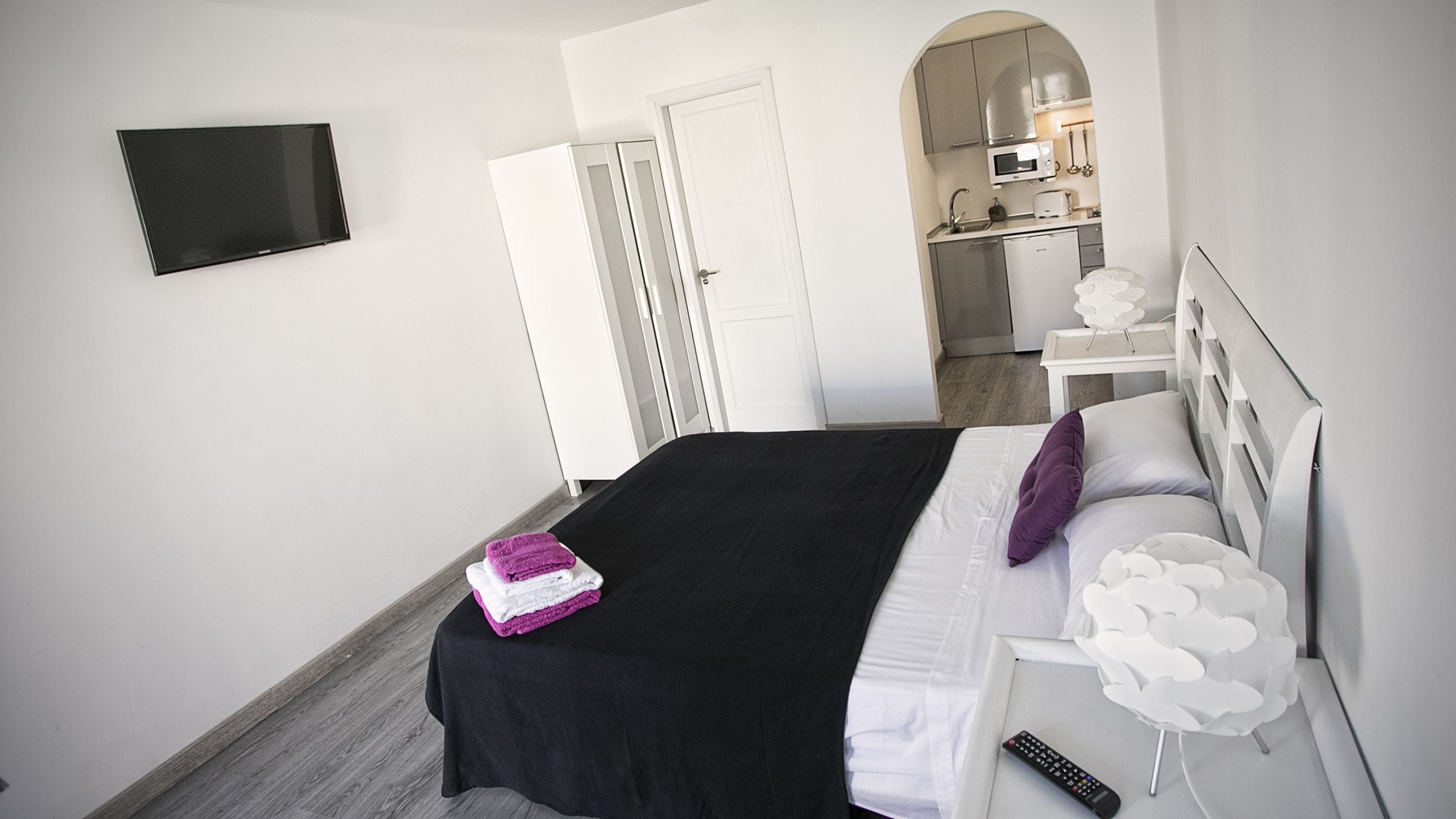 Une chambre d’hôtel avec un lit, un téléviseur fixé au mur, une petite kitchenette et une salle de bain visible à travers une arche.