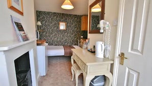 Une petite pièce, de taille modeste, comprenant un lit, un bureau, une chaise, une poubelle, une lampe, un miroir et un mur recouvert d’un papier peint à motifs.