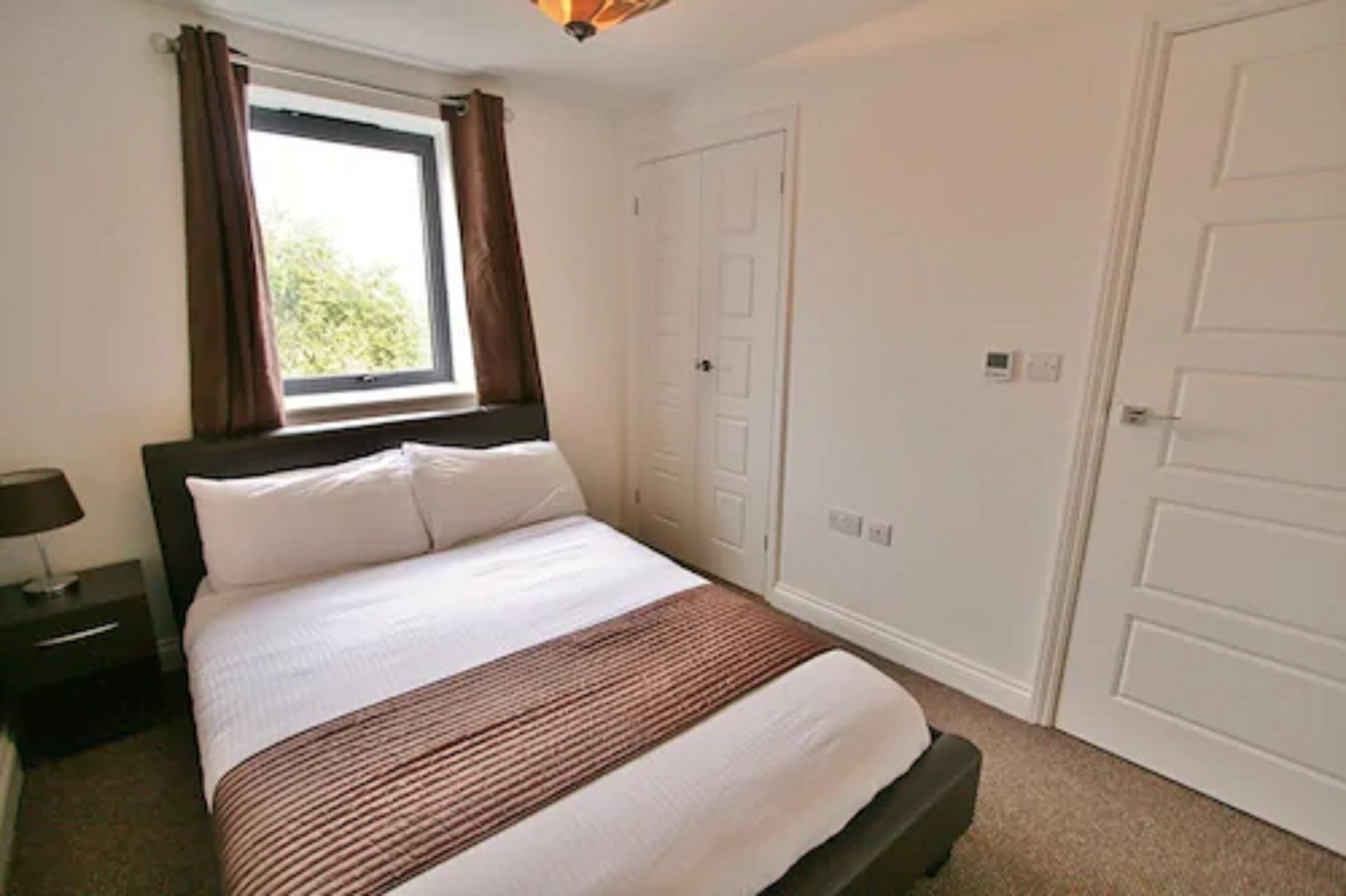 Un dormitorio con cama, mesita de noche, ventana con cortinas y puerta.