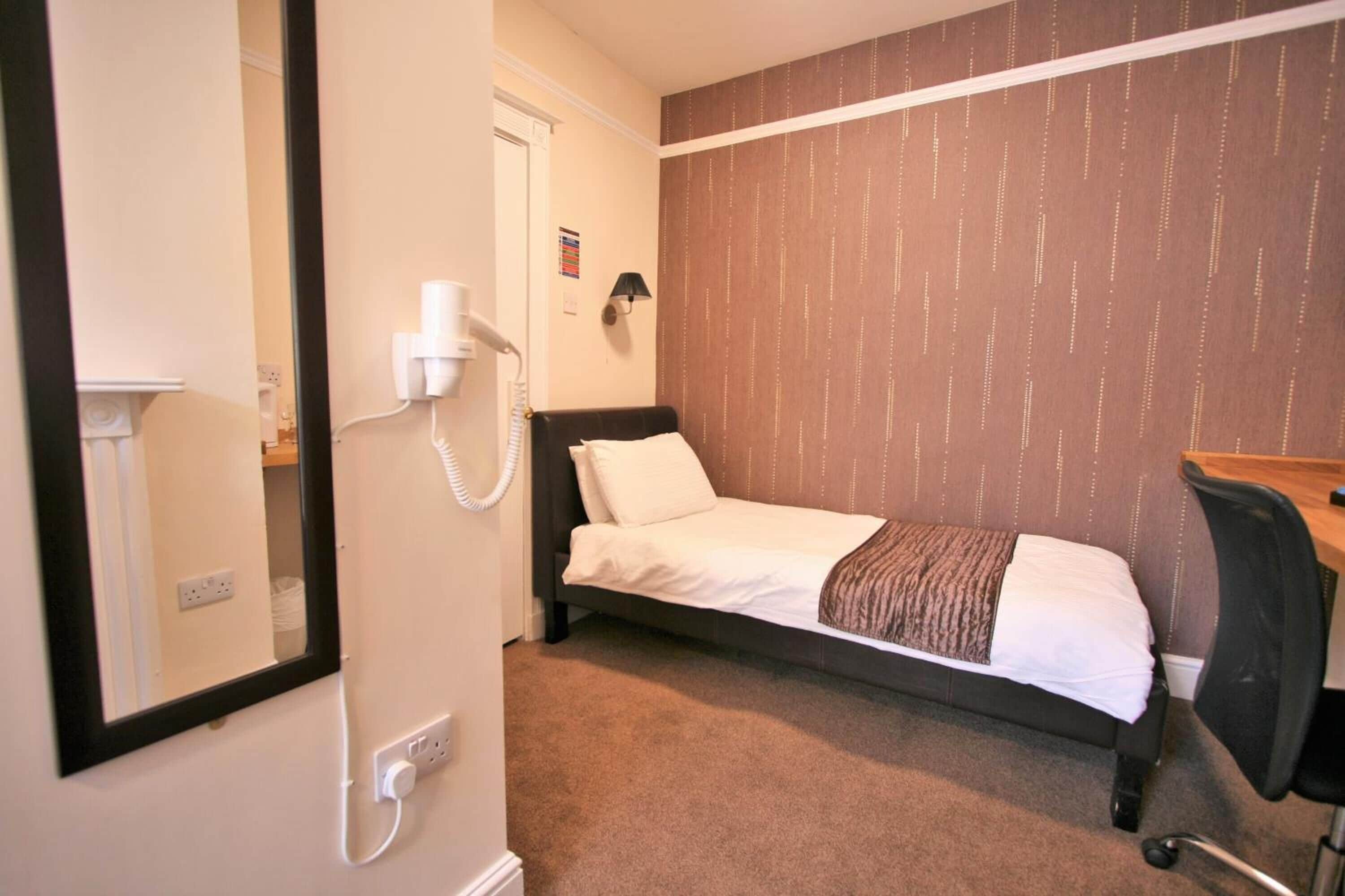 A small, neatly arranged hotel room with a single bed, a desk, and a chair. There is a telephone on the wall and a power outlet near the bed.
