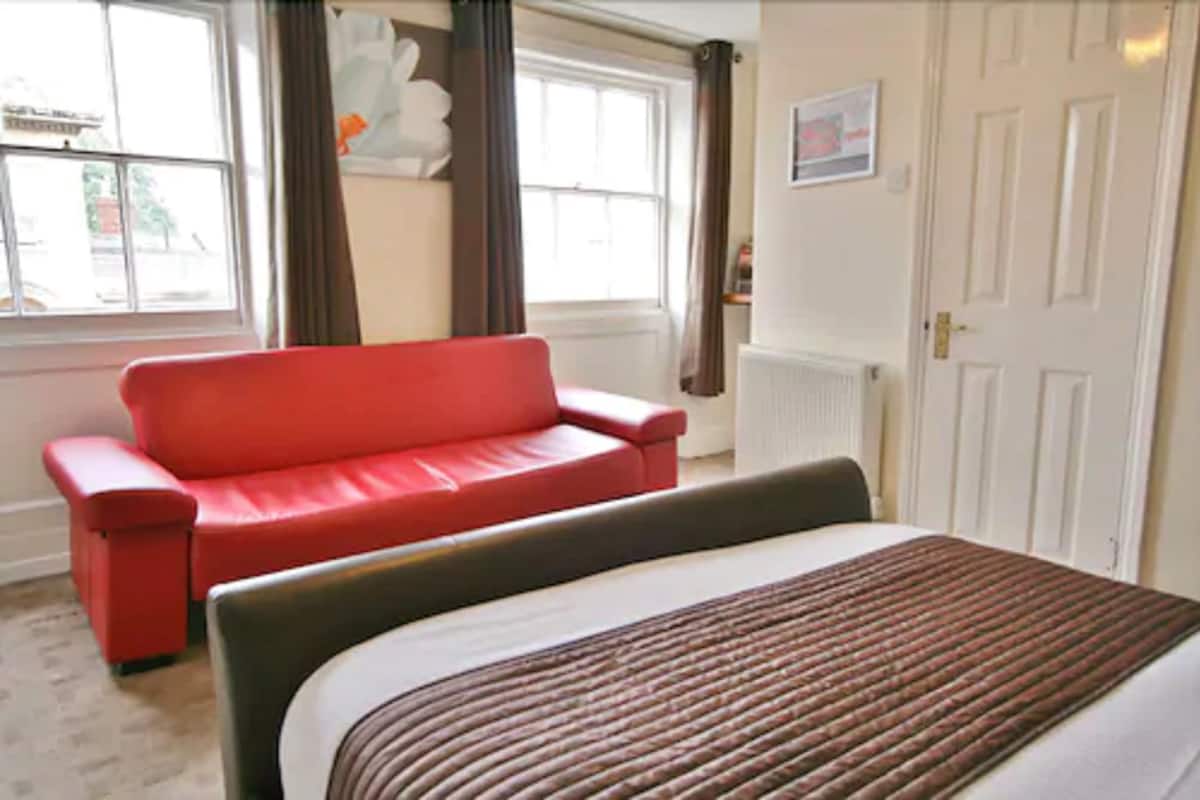 Ein Hotelzimmer mit einem roten Sofa, einem Bett mit gestreifter Bettdecke, einem Fenster mit Vorhängen und einer weißen Tür.