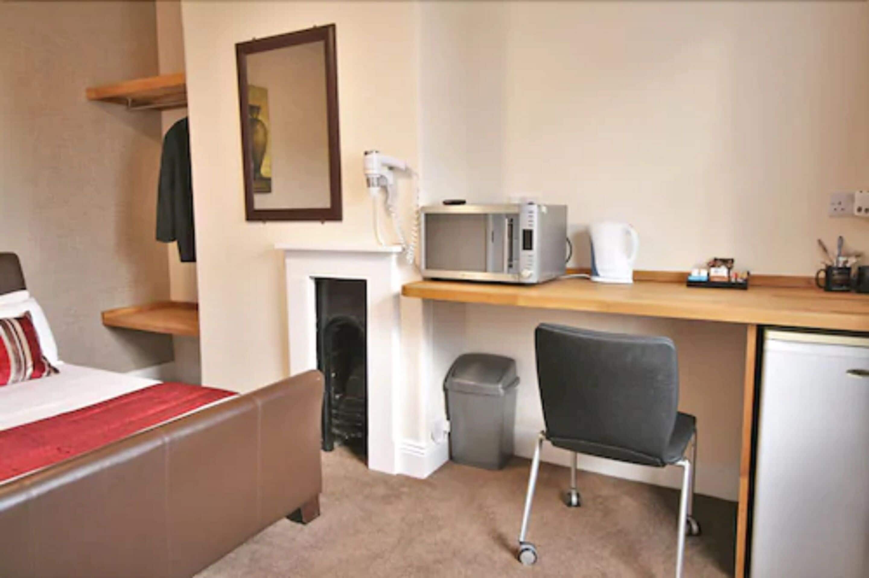 A hotel room with a bed, a desk with a microwave, a chair, a small trash bin, and a white wall with a framed picture.