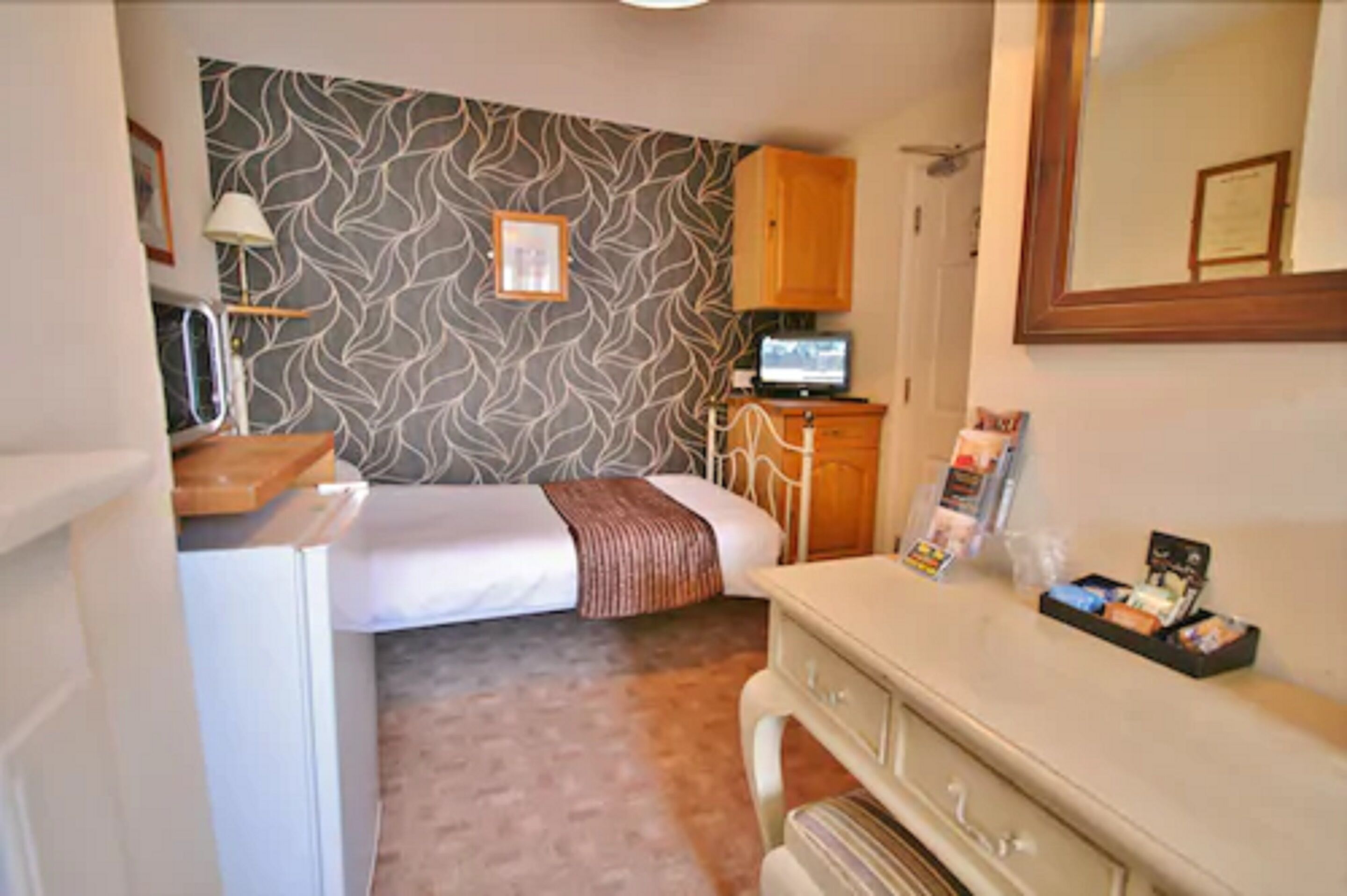 A compact hotel room with a bed, a dresser, a mirror, a television, and a patterned wall.