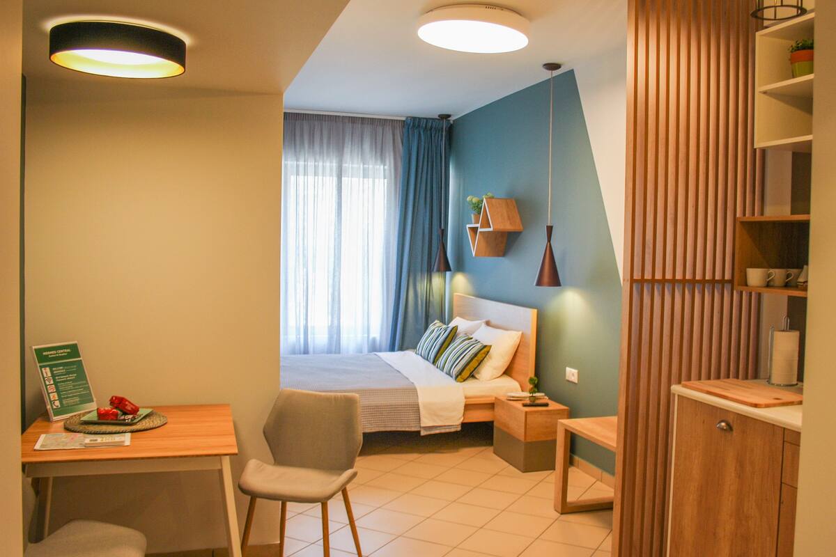 A modern hotel room with a bed, a desk, a chair, a nightstand, and a wooden wardrobe.
